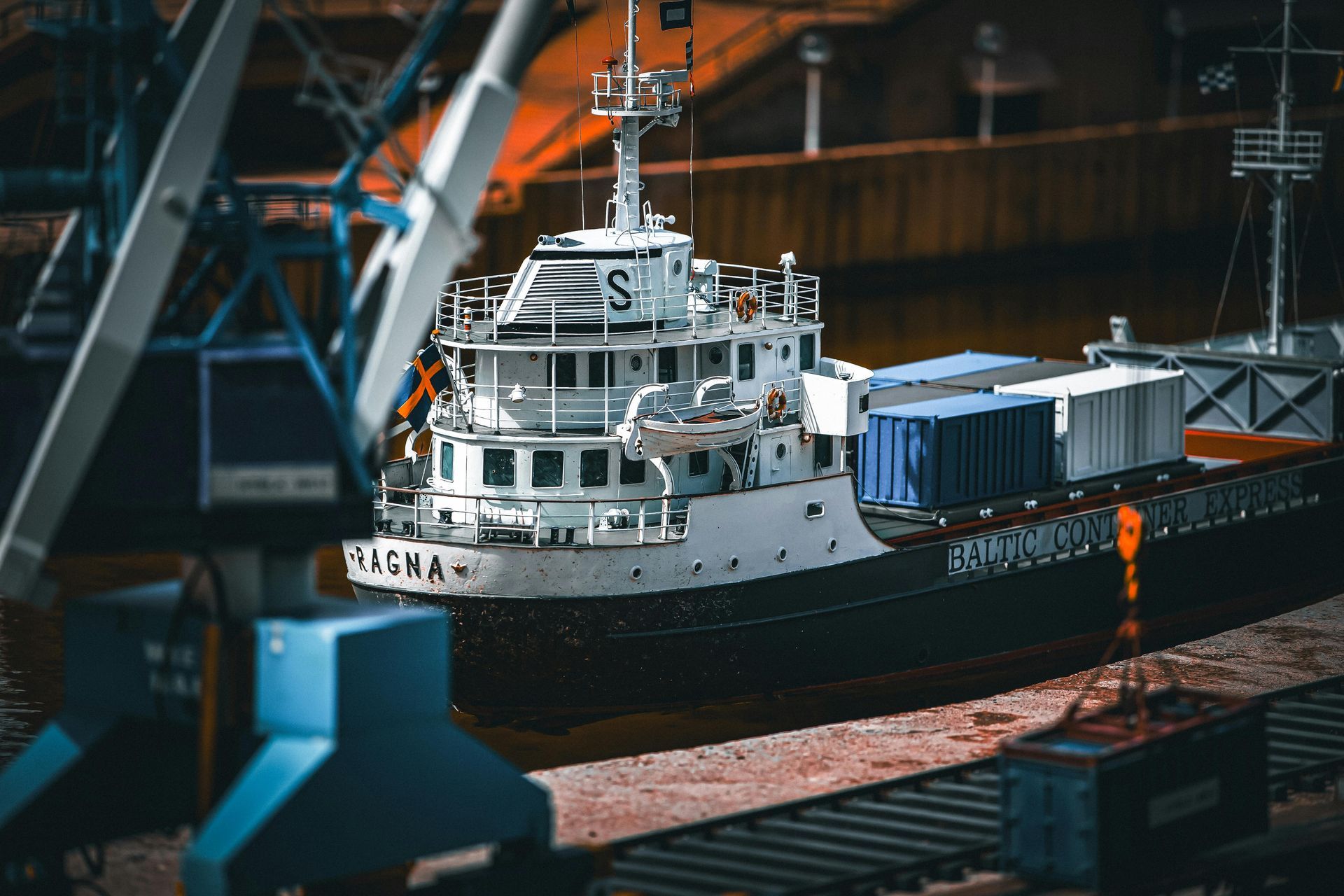 A detailed scale model of a cargo ship with blue and white containers, docked beside a blue crane and railroad tracks.