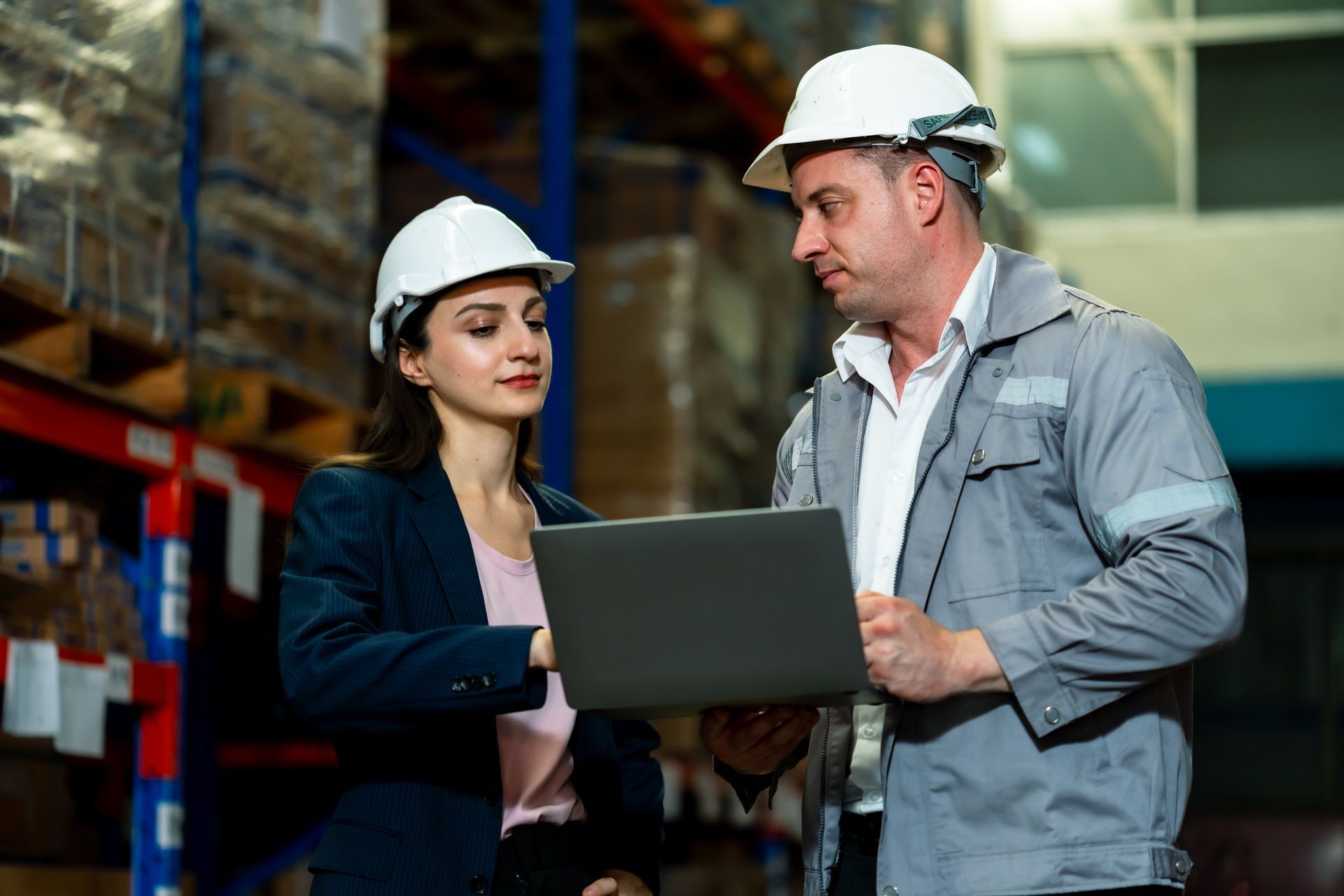 A woman in a blazer and a man in work coveralls, both wearing hard hats, review information on a laptop in a warehouse.