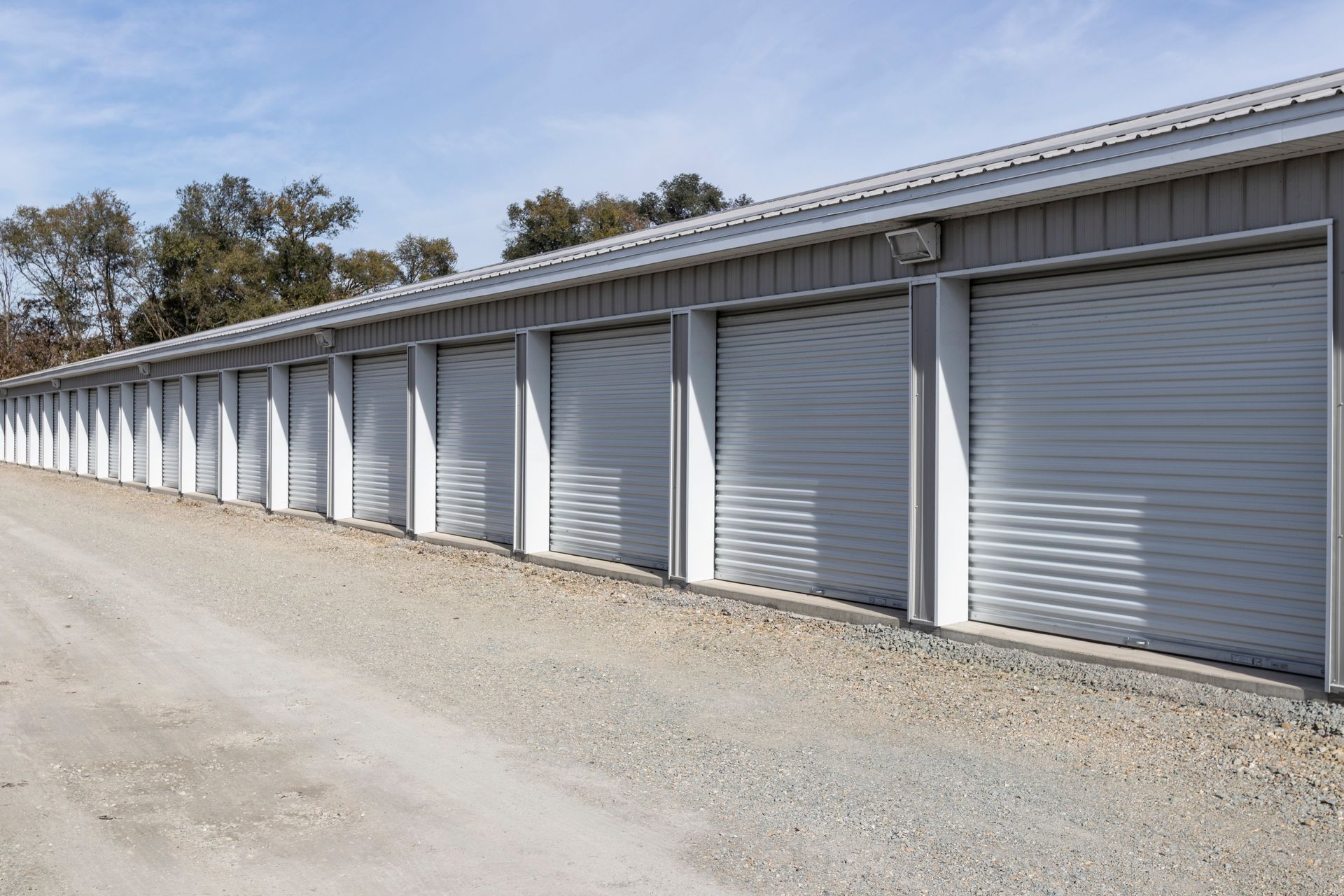 A long row of storage units with closed roll up doors along a gravel driveway. A long row of storage units with closed roll up doors along a gravel driveway.