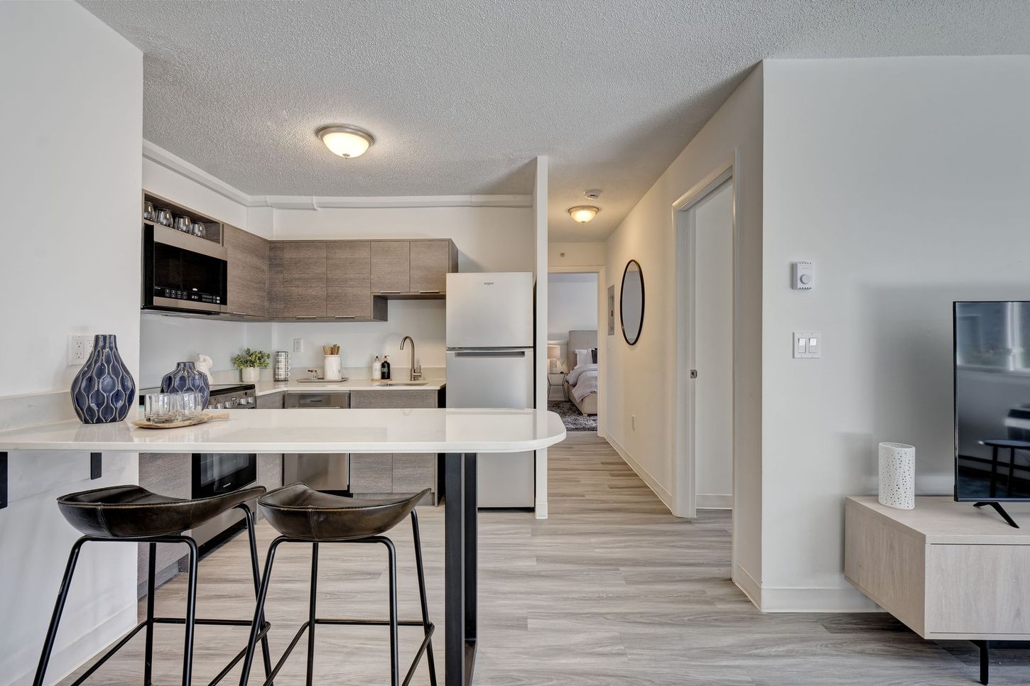 Interior view of an apartment with a kitchen, living area, and hallway; features neutral tones, bar stools, and modern appliances.