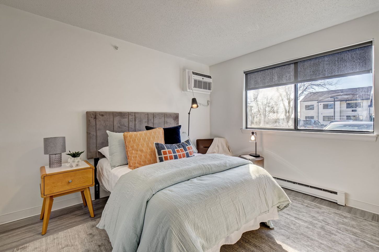 Bedroom with bed, nightstands, and a window. Light blue bedding with colorful pillows, light gray walls.