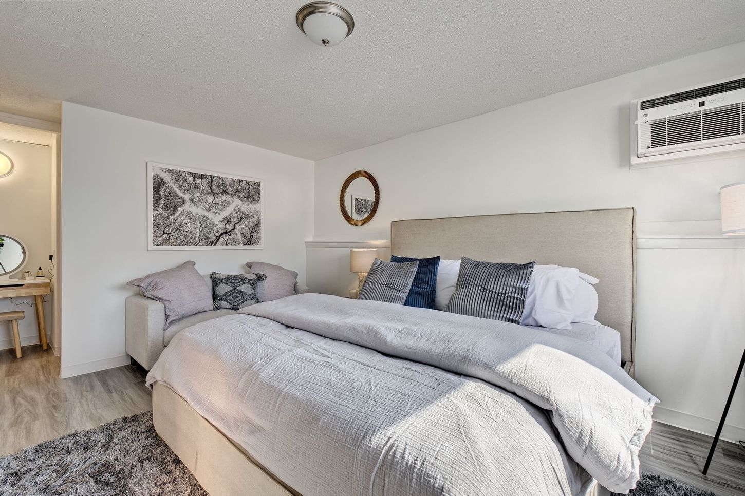 Bedroom with bed, small couch, art, mirror, and air conditioner. White walls and light wood floor.