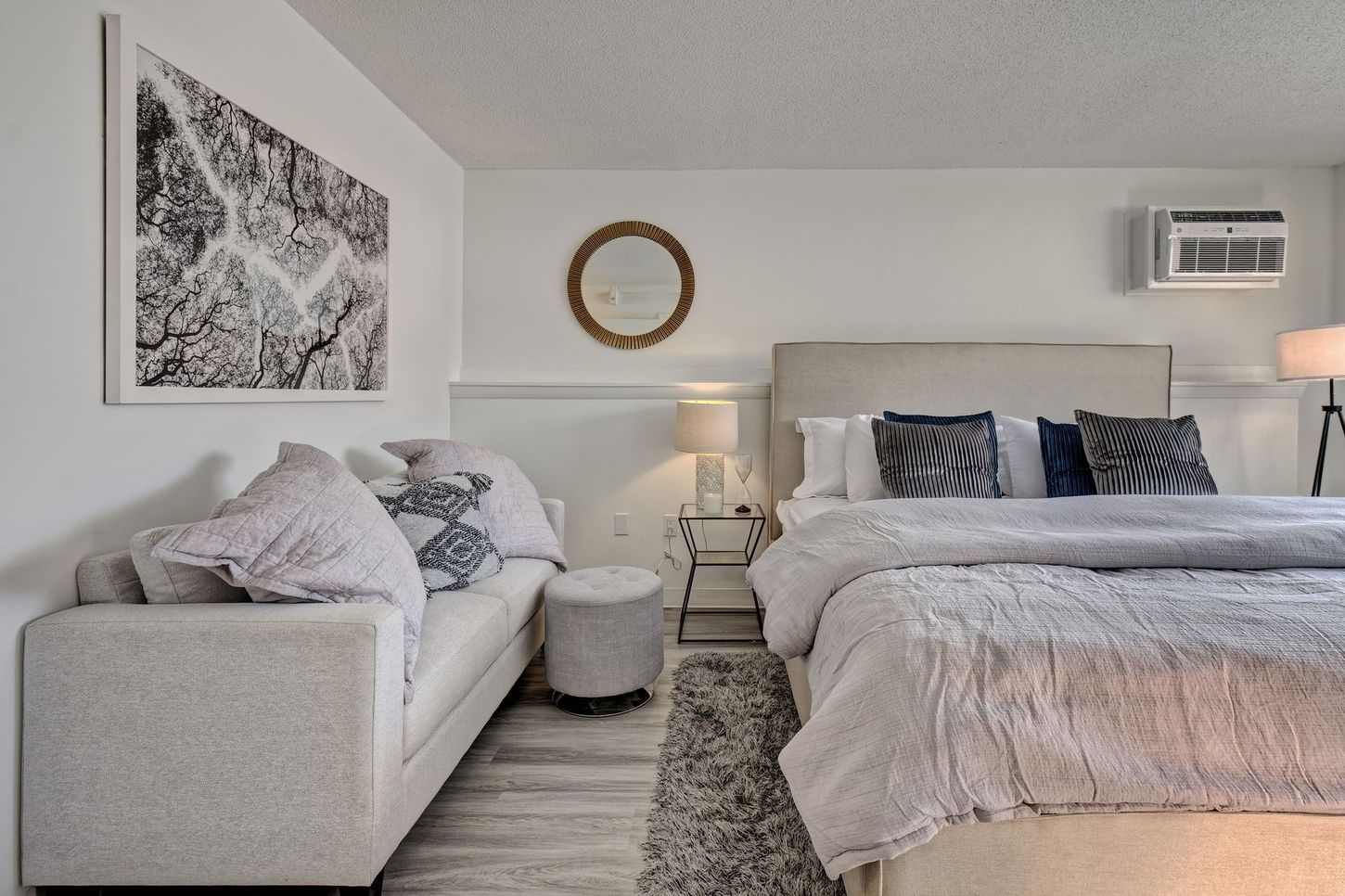 White studio apartment with bed, couch, artwork, and air conditioner.