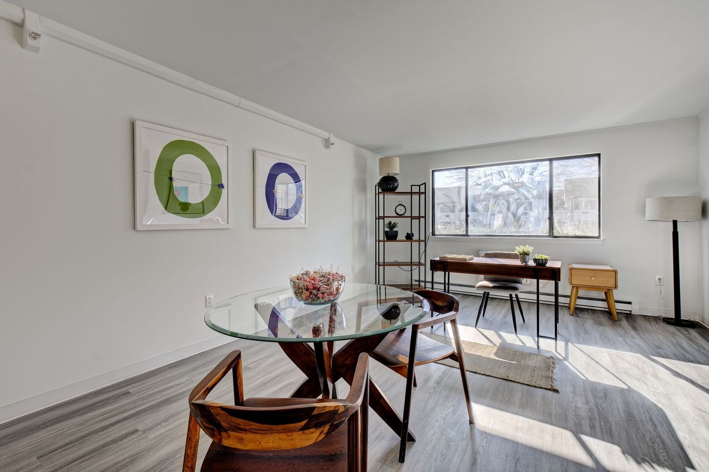 A bright room features a glass-top dining table, artwork, a desk, and a bookshelf near a large window.