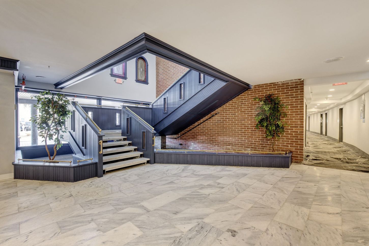 Lobby with stairs, marble floor, brick wall, and decorative plants. Hallway with doors in the background.