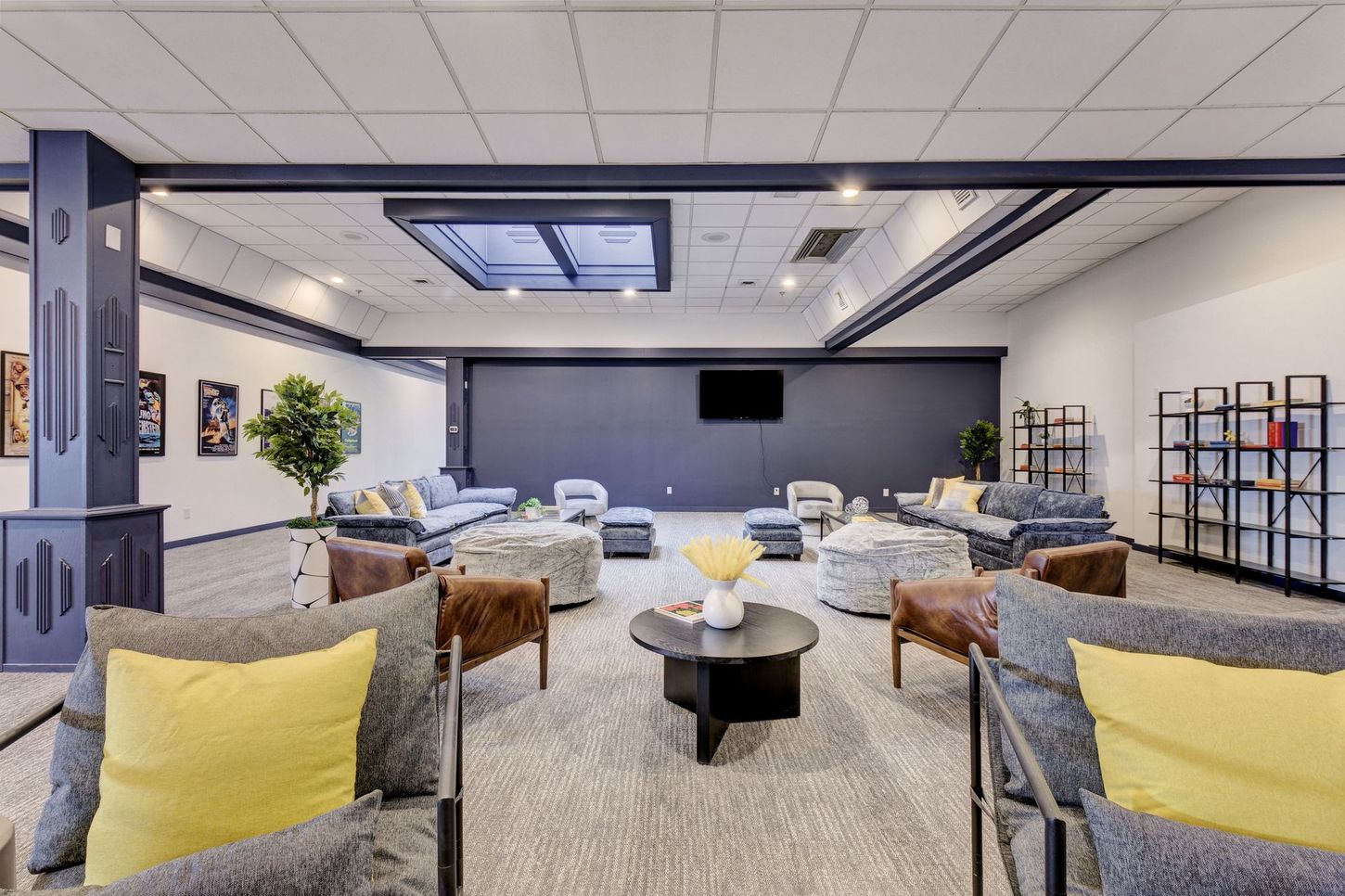 Spacious lounge with couches, ottomans, bookshelves, and a TV on a dark wall. Neutral tones with yellow accents.