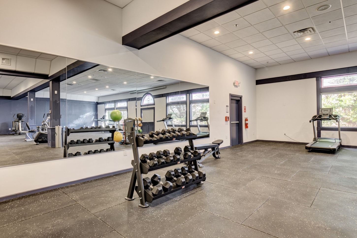 Gym with weights, treadmill, and large mirror. Gray and white walls, dark flooring.