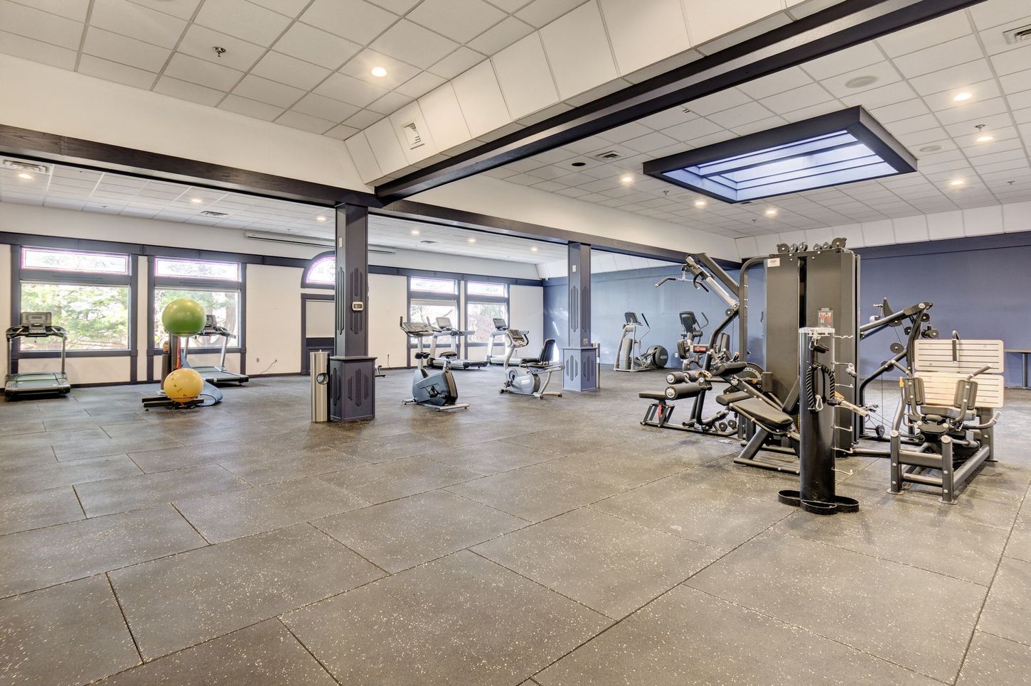 Gym with workout equipment on a rubber floor; large windows and bright ceiling lights.