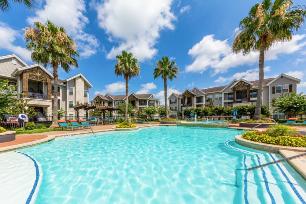 A large swimming pool surrounded by palm trees in front of apartment buildings at Marquis at Sugar Land in Sugar Land, TX.