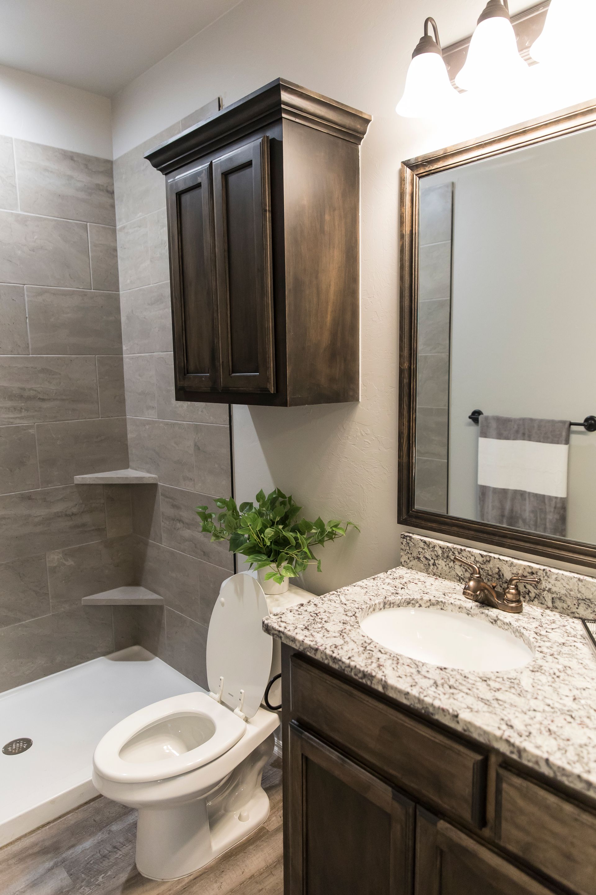 Bathroom with gray tile shower, brown vanity and cabinet, toilet, and white sink.