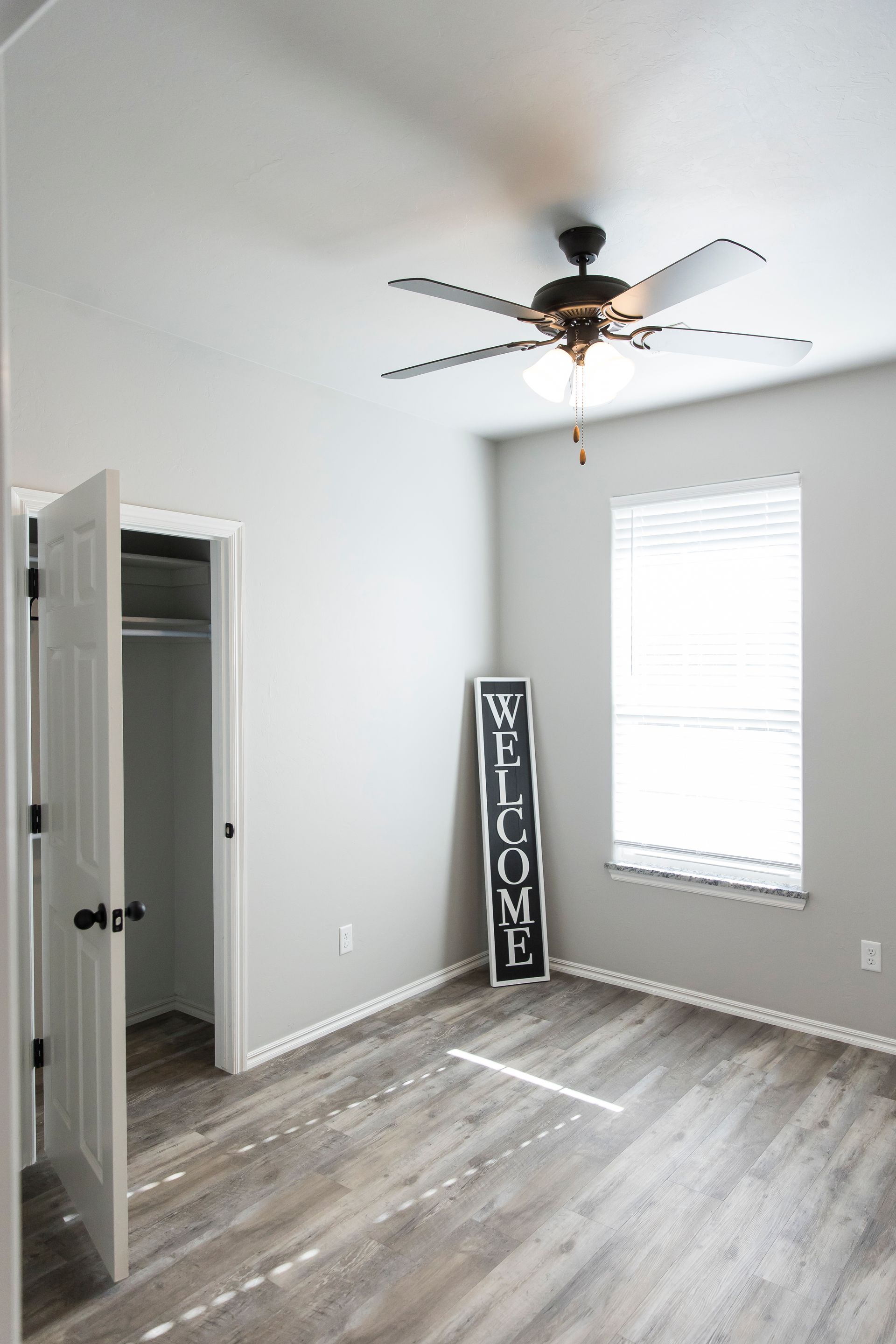 Empty bedroom with gray walls, wooden floor, and closet. A Welcome sign stands near a window.