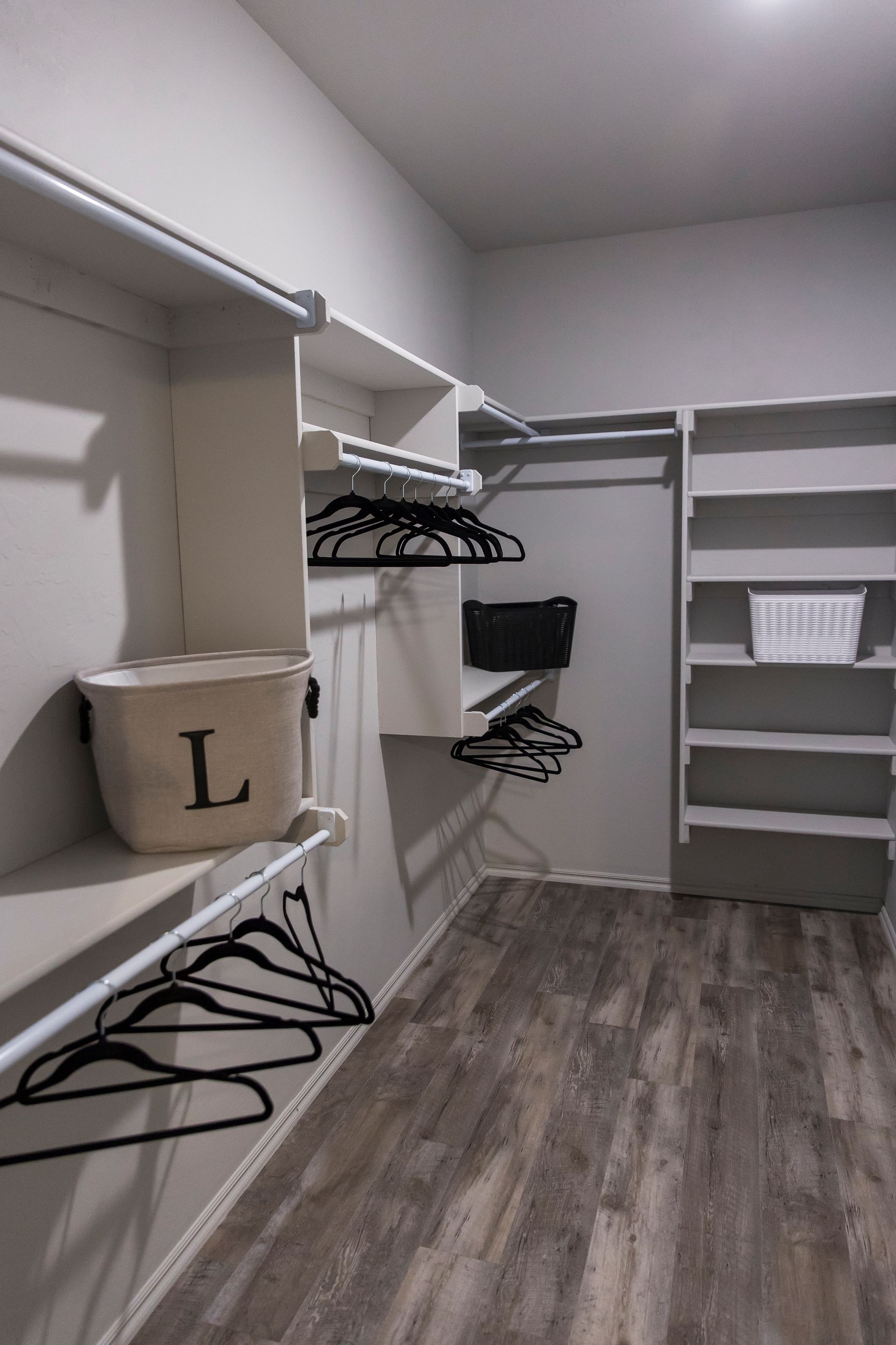A walk-in closet with white shelves, rods, and wooden floor, holding black hangers and a laundry basket.