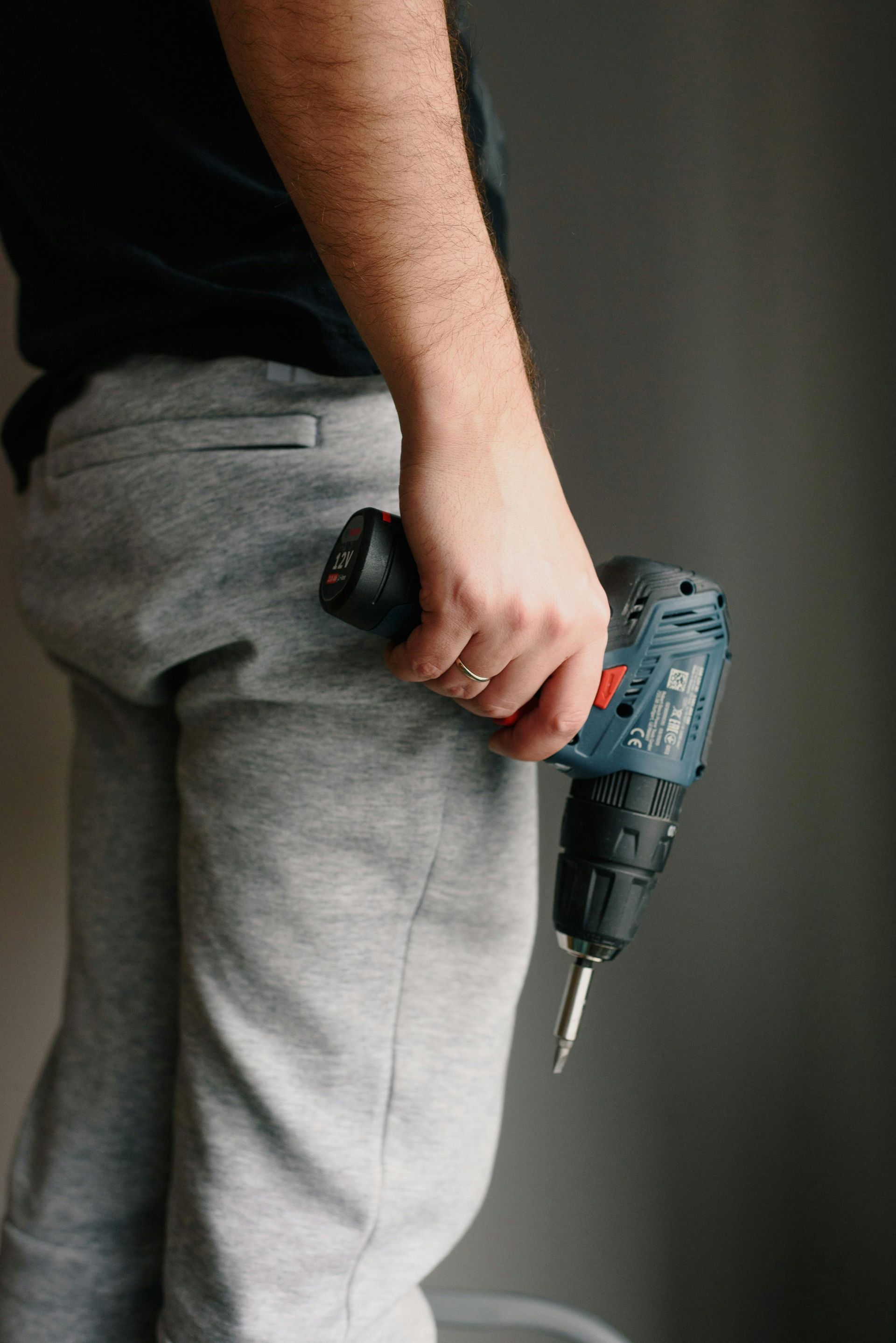Person holding a cordless drill, wearing gray sweatpants and a black shirt, indoors.