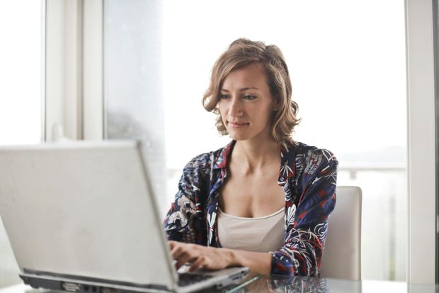 Woman with wavy hair sits at table, typing on laptop in bright room.