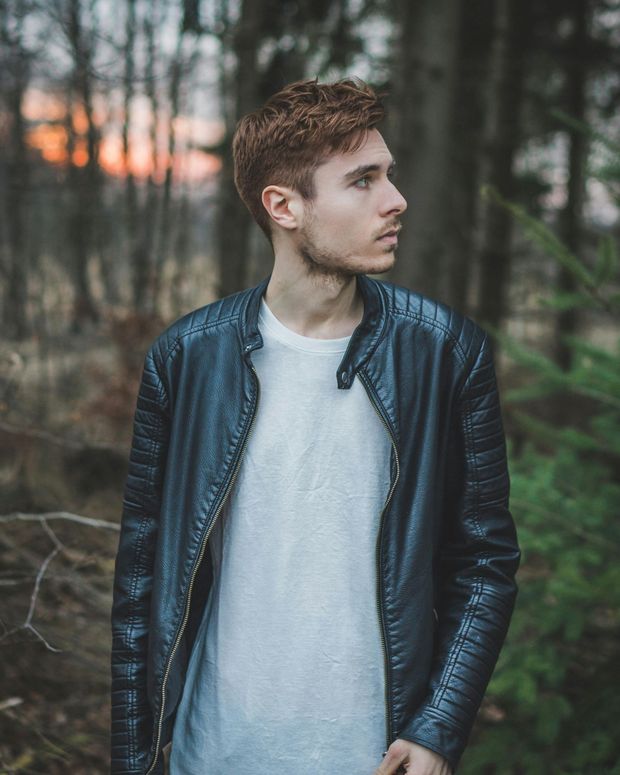 Young man with red hair wearing a black leather jacket and white shirt, looking off to the side in a forest.