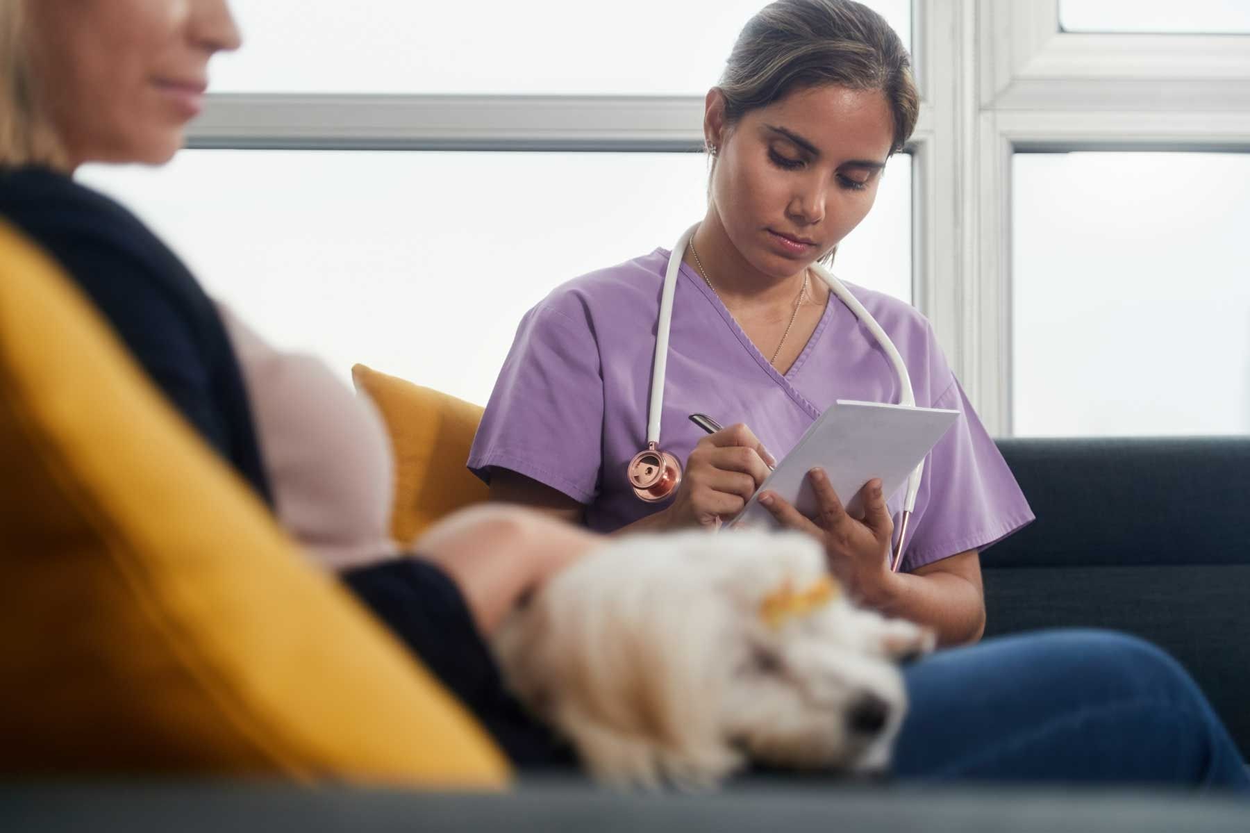 A Woman Is Sitting On A Couch With A Dog While A Nurse Writes On A Clipboard — Ballina Veterinary Hospital In Ballina, NSW