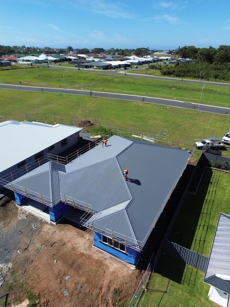 Aerial View of a House With a Dark Grey Roof, Two Workers on the Roof, and a Green Field — Ace1 Metal Roofing in Forster, NSW
