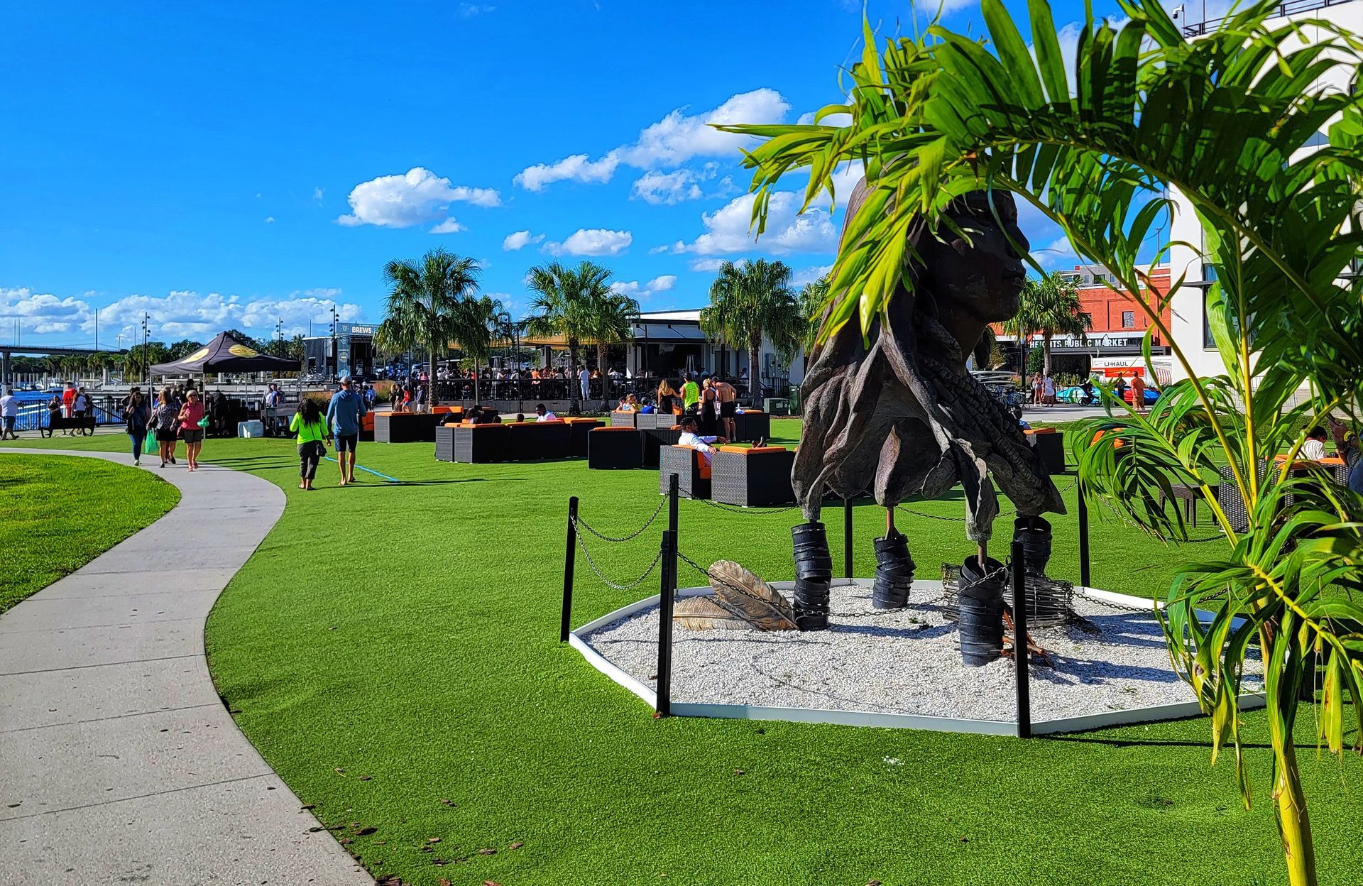 A park with green grass, a paved path, and an abstract sculpture; people are visible in the background.