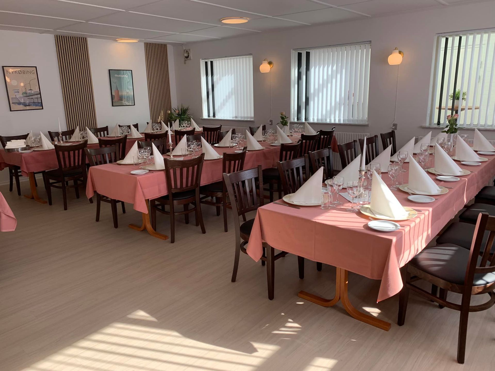 Spisestue med lyserøde duge, dækkede borde og stole samt et stort vindue.