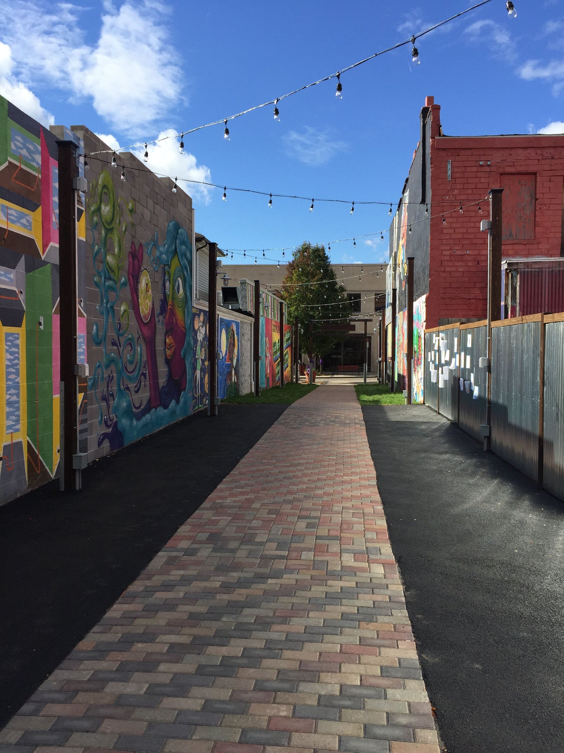 Brick walkway between buildings with murals, string lights overhead, under a blue sky.
