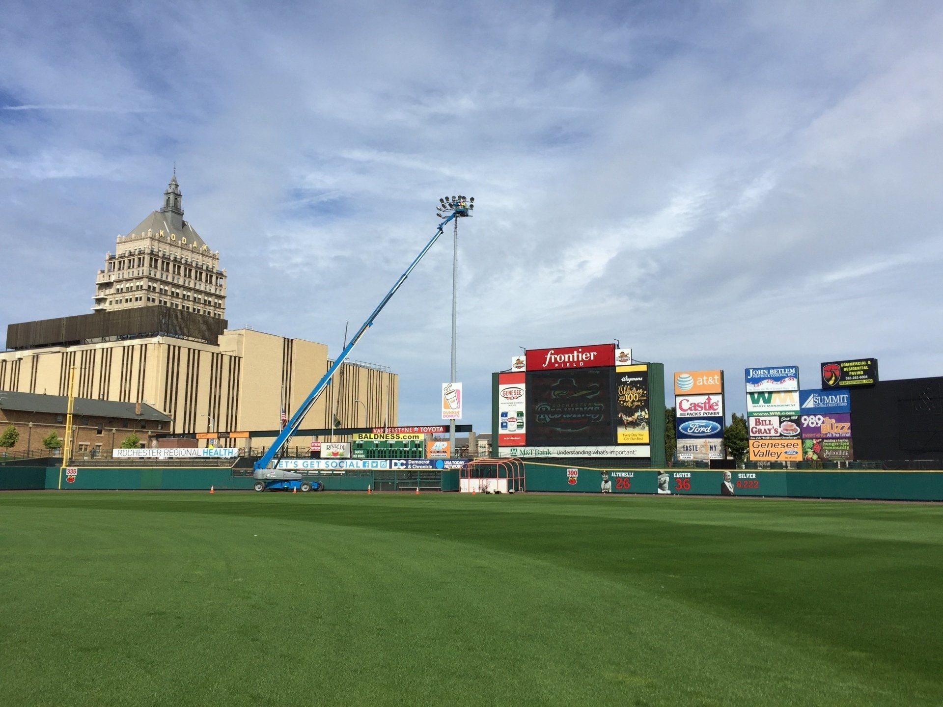Baseball field with green grass, scoreboard, blue lift, and large building in background.
