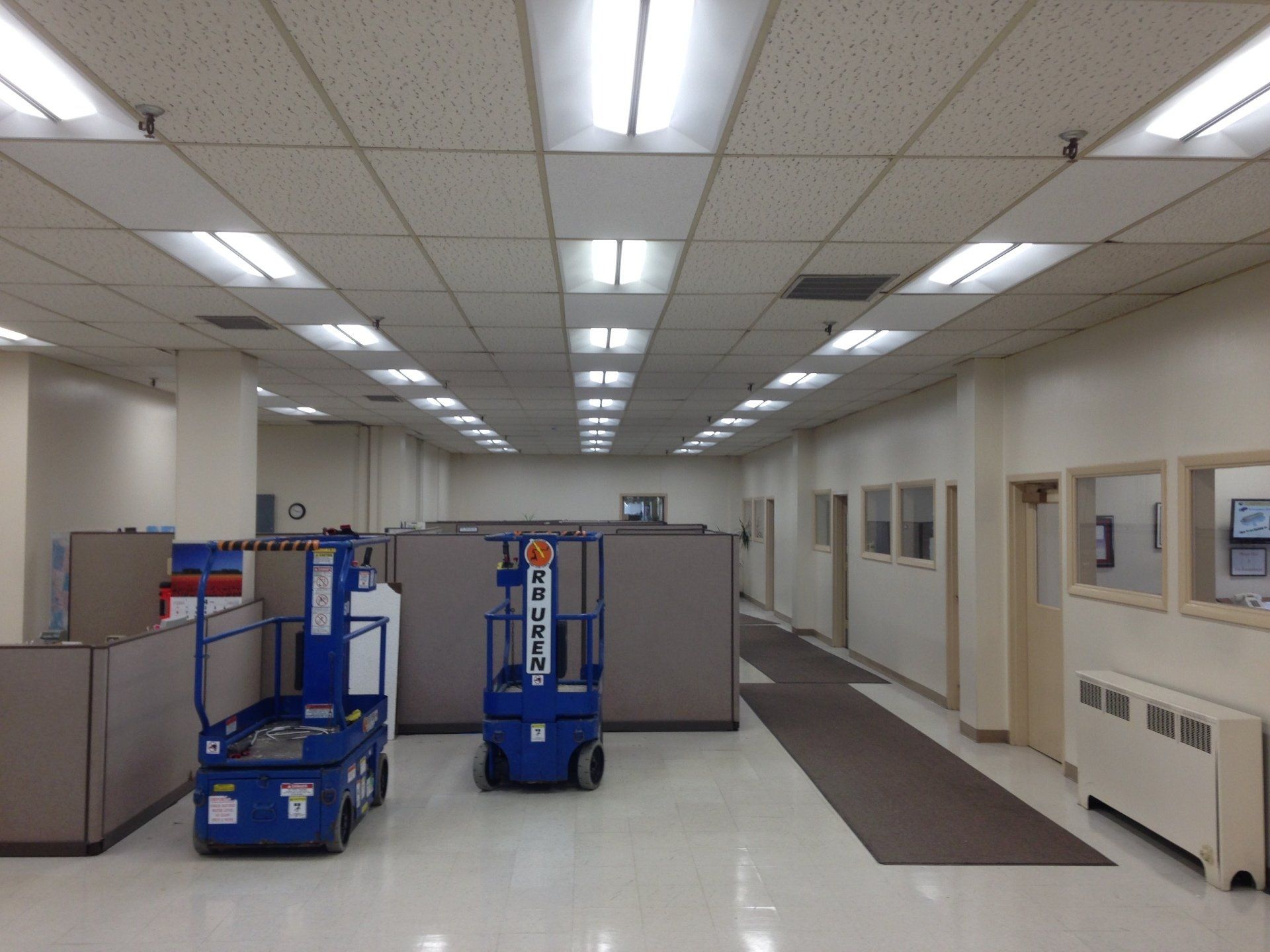 An interior office with cubicles, a hallway, and two blue lift machines under bright fluorescent lights.