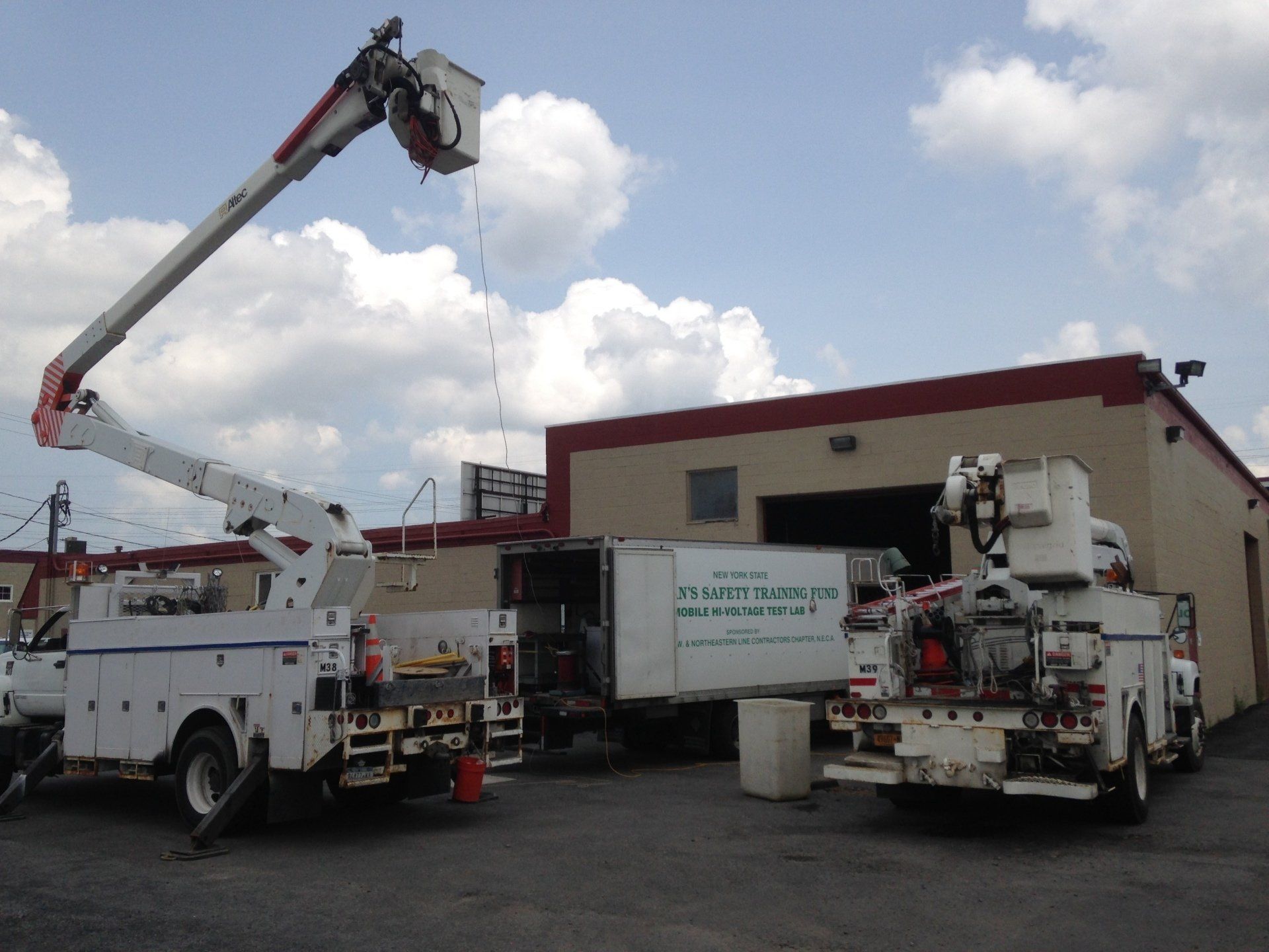 Two white bucket trucks with booms extended near a tan building. Blue sky and clouds.