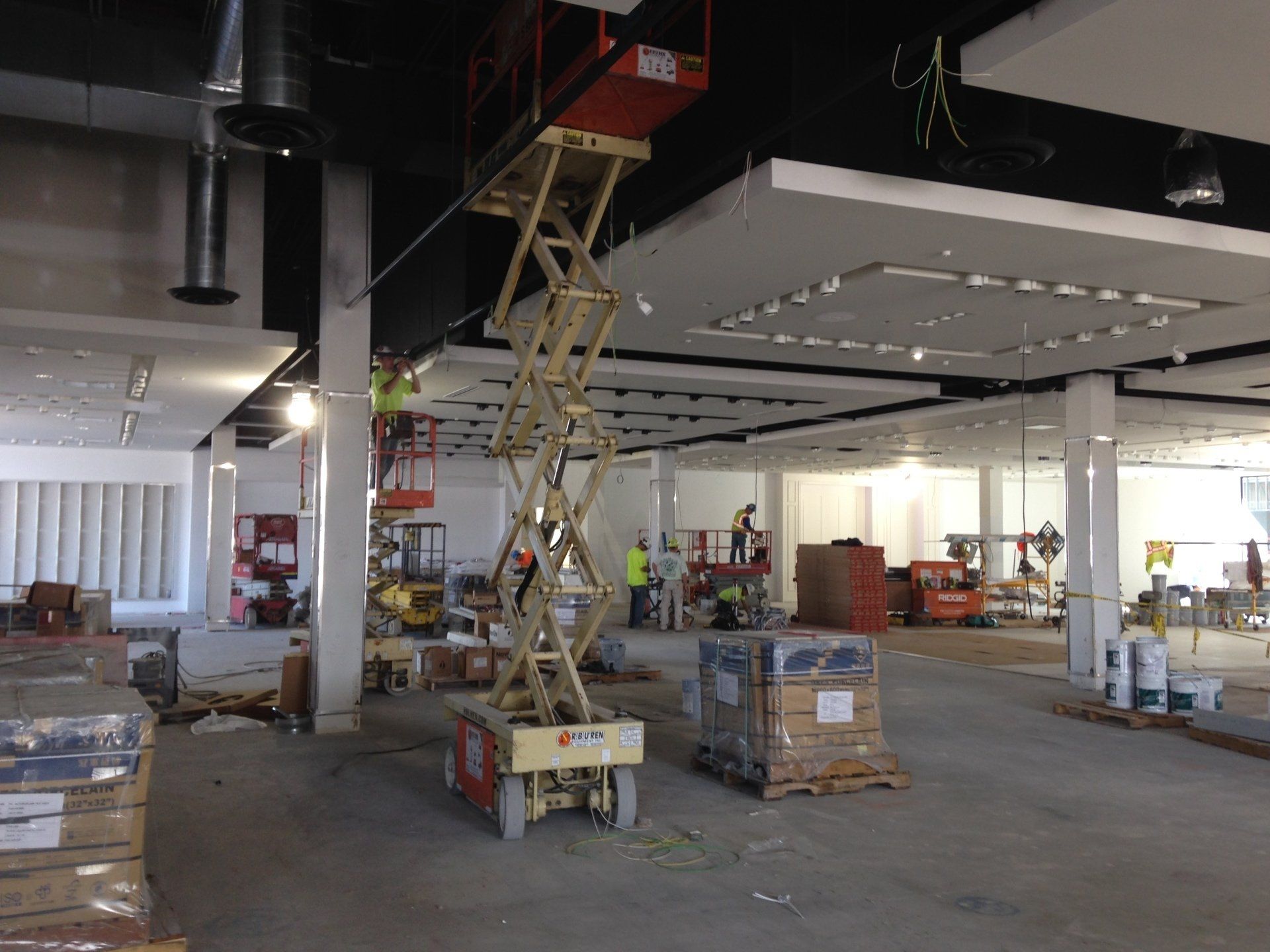 Construction site: workers on lifts installing ceiling lights, pallets of materials on the floor.