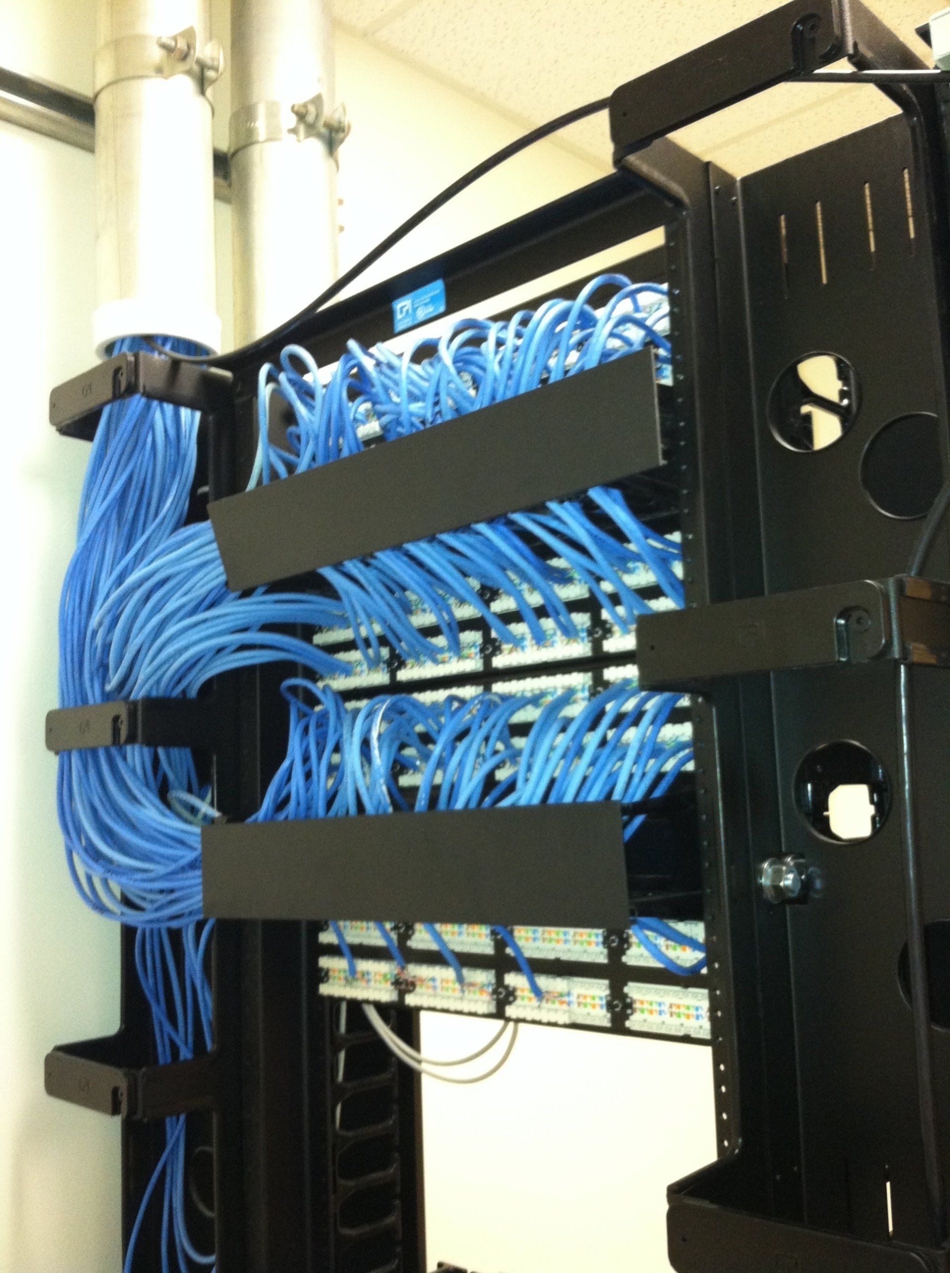 Blue network cables organized in a black rack; white pipes are visible in the background.
