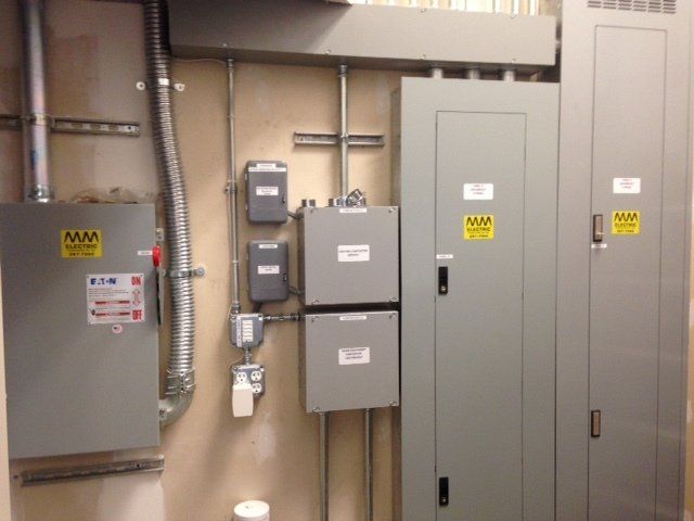 Electrical panel and components on a wall, including gray metal boxes, conduits, and safety labels.