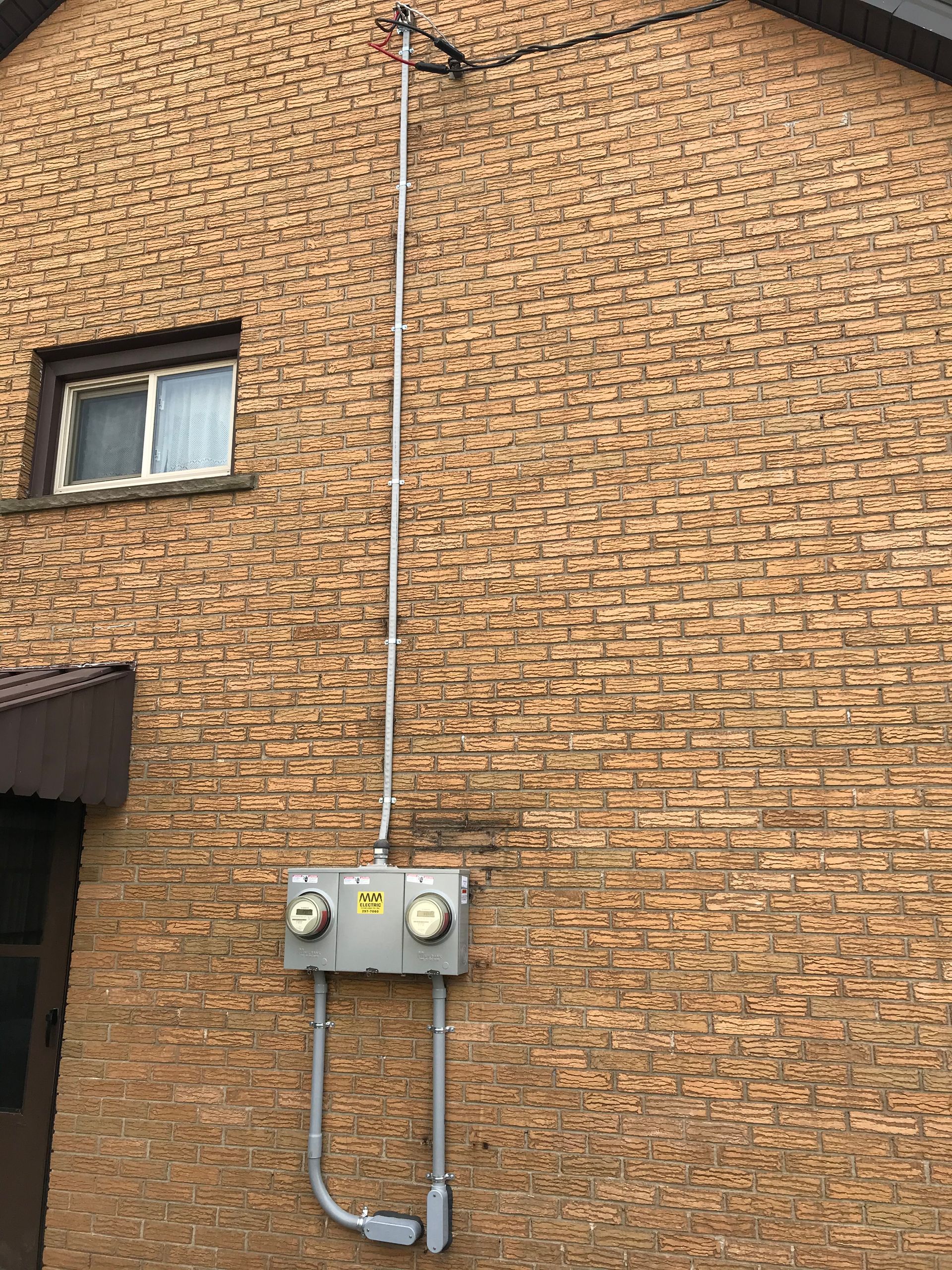 Electrical meter box on a brick wall with conduit leading to an overhead wire.