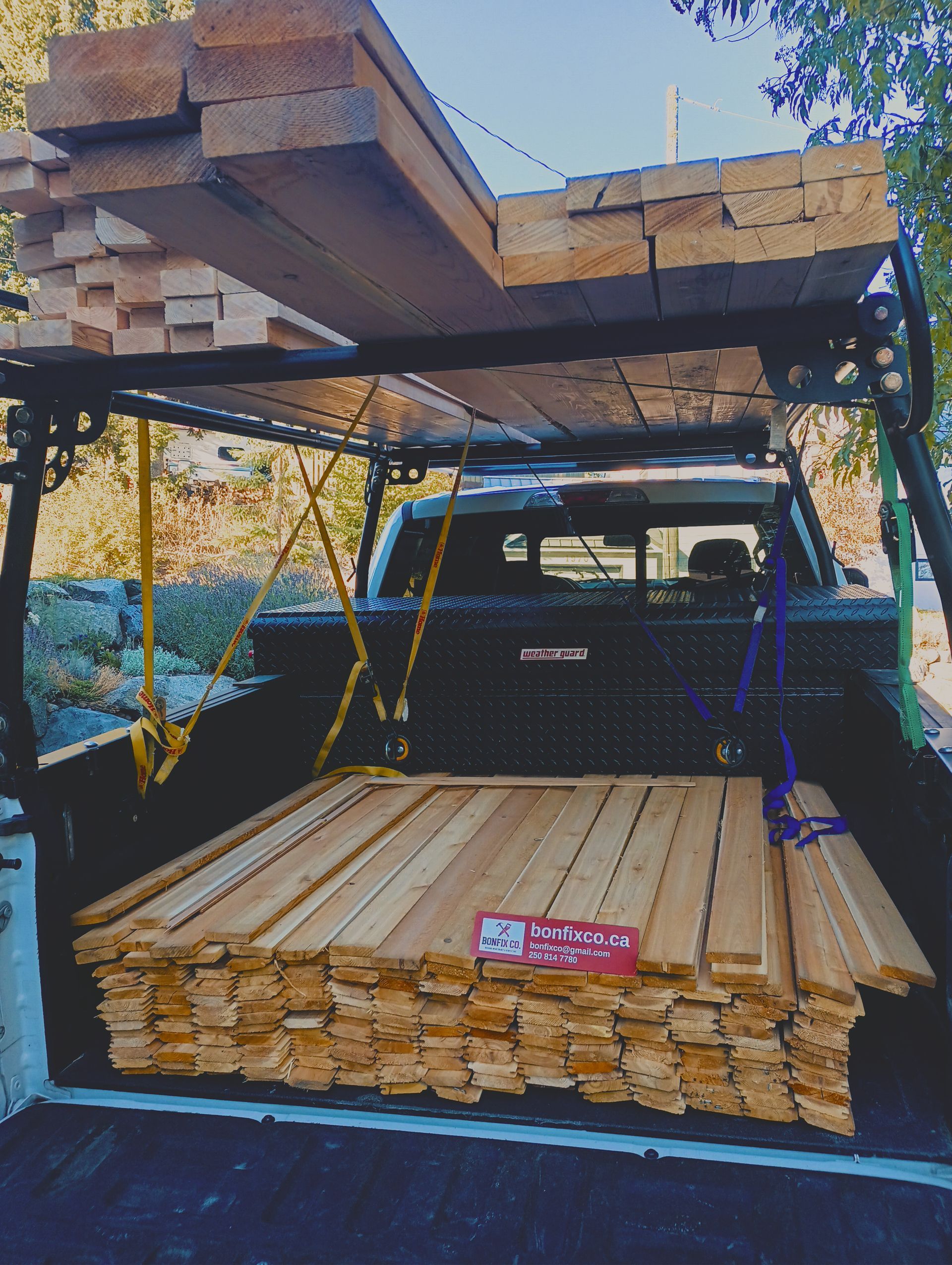 A pickup truck bed loaded with stacks of lumber, with more wooden boards secured to a metal rack overhead.