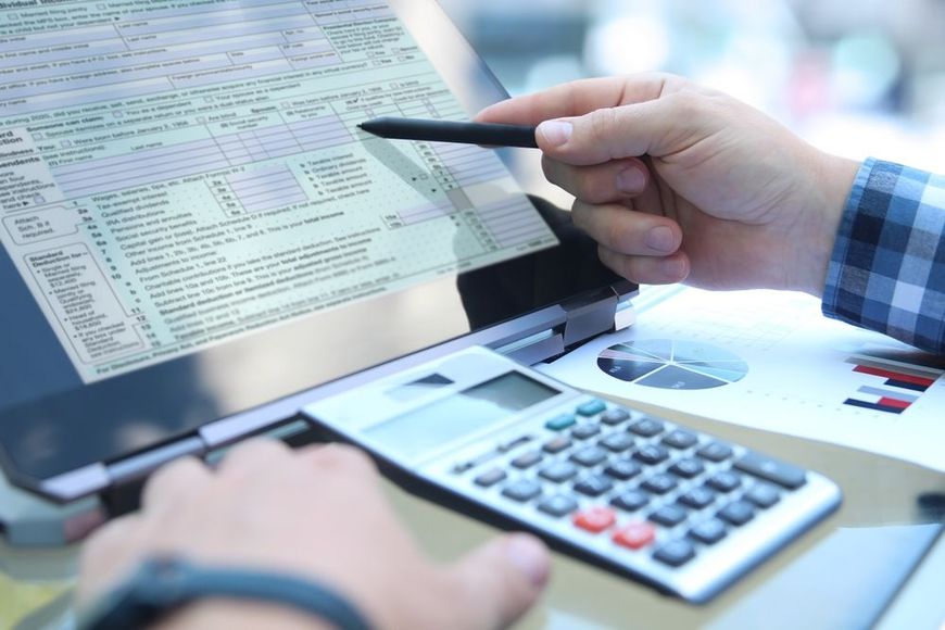 Person using a stylus to point at a form on a computer screen, with a calculator and graphs on a desk.