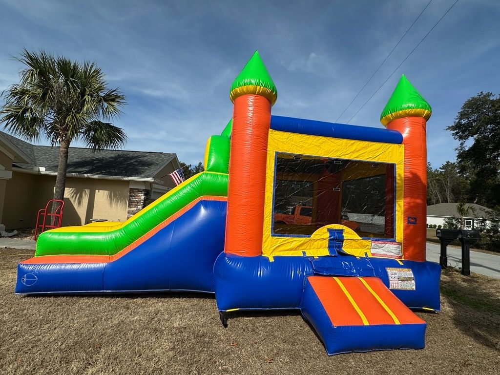 A colorful, inflatable bounce house with a slide on the left, set on a grassy lawn in front of a residential home.