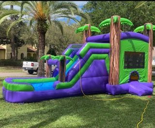 A purple and green inflatable bounce house with a slide and palm tree accents set up on a grassy lawn.