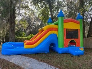A colorful inflatable bounce house with a slide and a shallow pool at the base, set outdoors on a grassy lawn.
