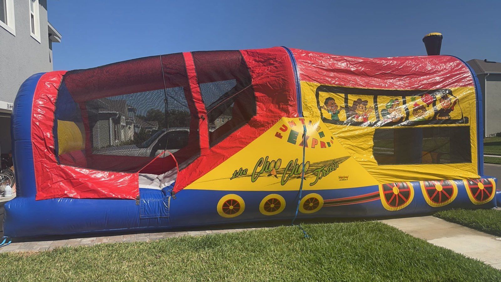A large, inflatable train-shaped bounce house in red, yellow, and blue sitting on a lawn.