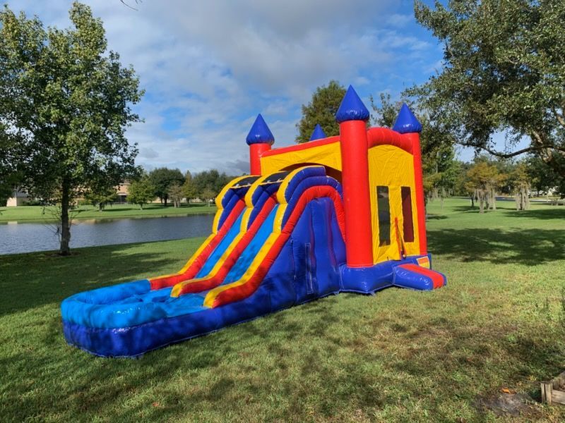 A vibrant red, yellow, and blue inflatable bounce house with a double-lane water slide on a grassy lawn by a lake.