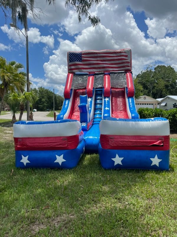 An inflatable dual-lane water slide with red, white, and blue stars-and-stripes decor set on a sunny grassy lawn.