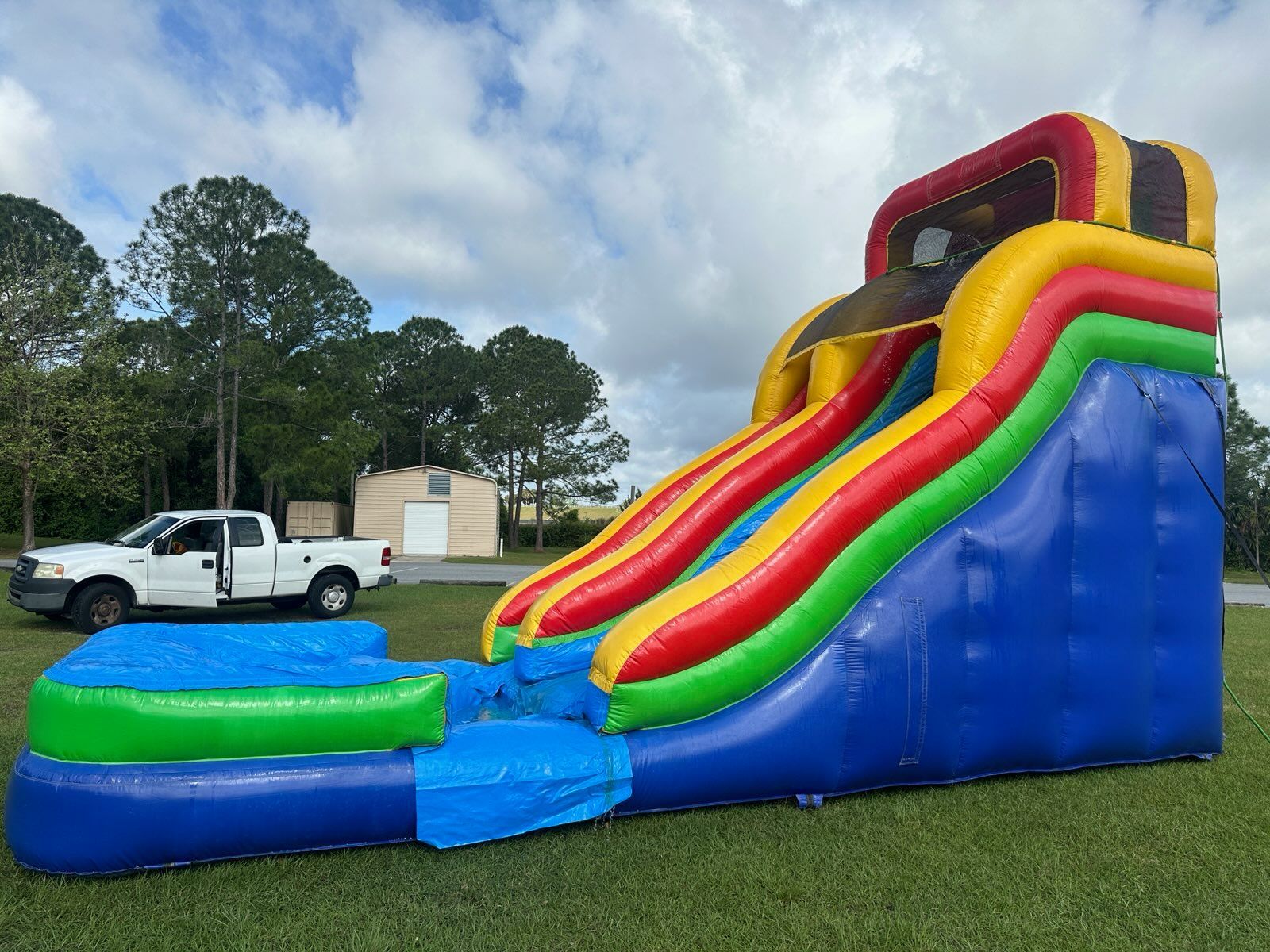A large, colorful inflatable slide in a grassy field with a white pickup truck parked in the background.