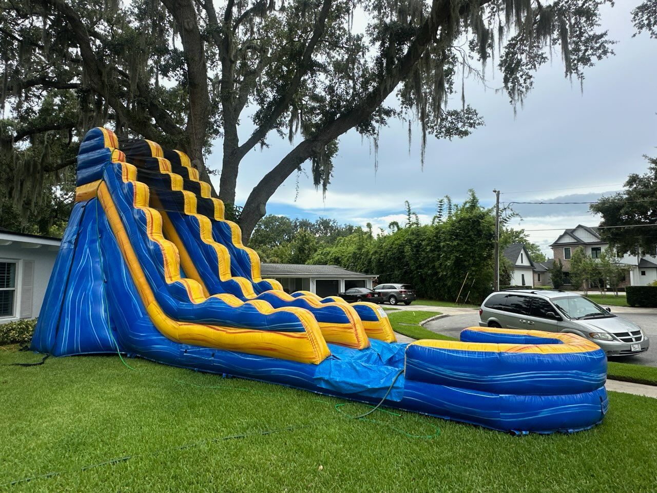 A large, blue and yellow inflatable water slide sits on a residential lawn under trees, with a car parked in the street.