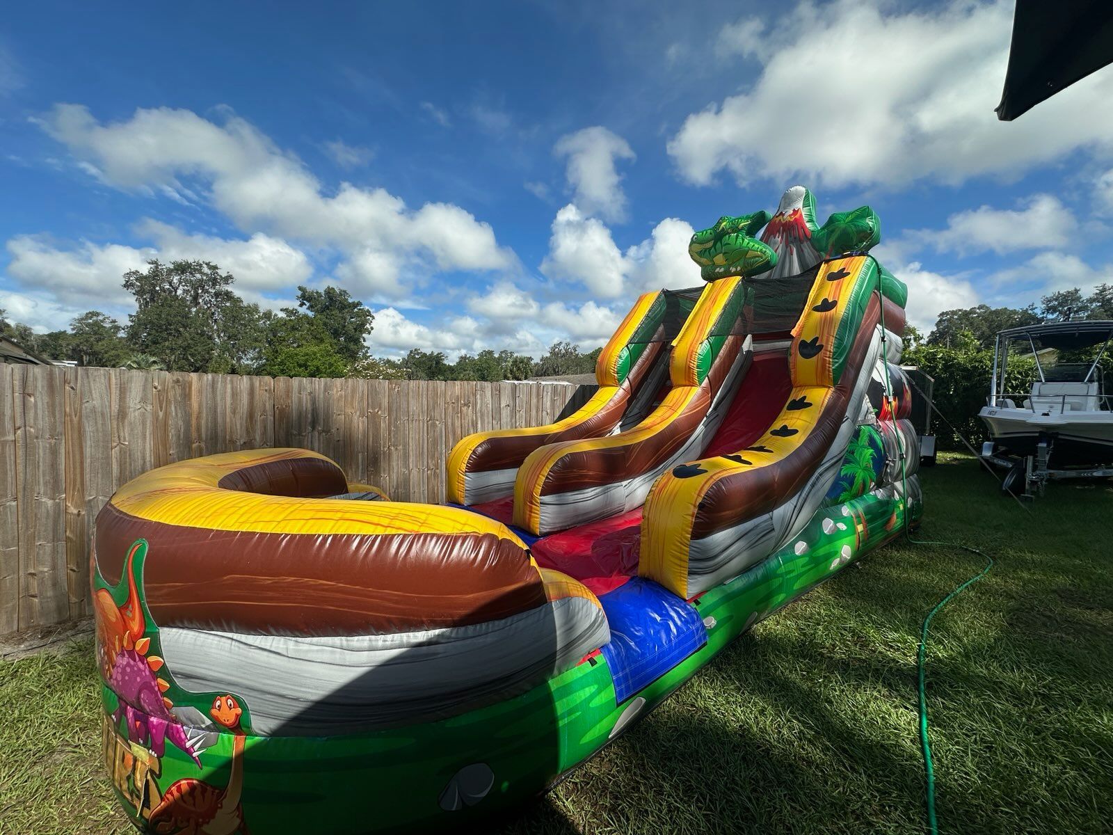 A dinosaur-themed inflatable water slide set up on a grassy lawn in front of a wooden fence.