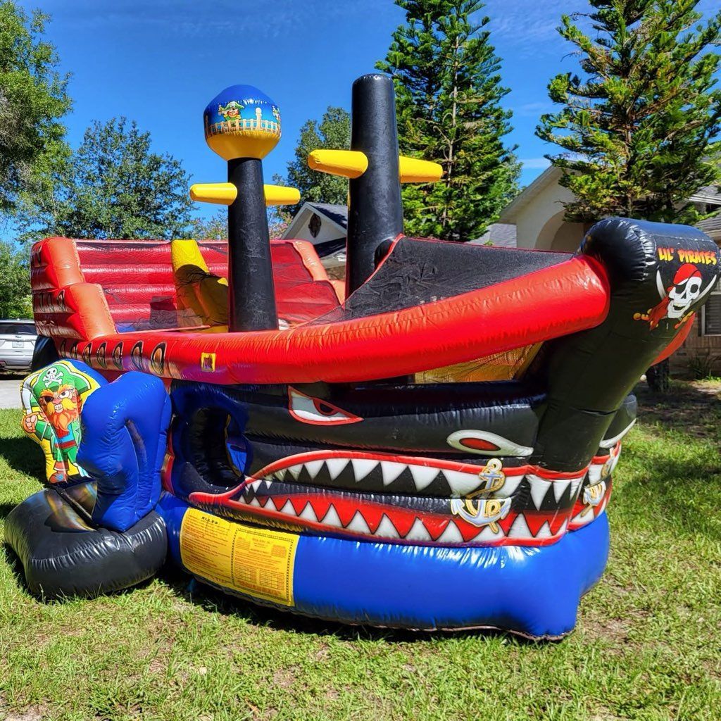 A colorful, pirate ship-themed inflatable bounce house sits on a green lawn in a sunny outdoor setting.
