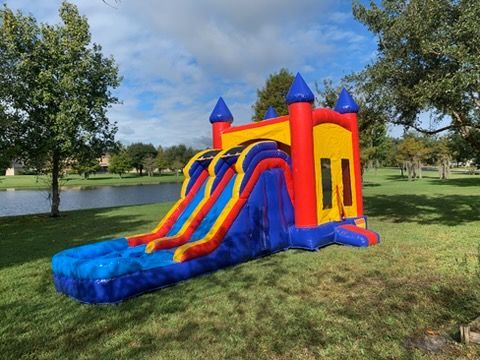 A red, yellow, and blue inflatable bounce house with a double water slide, set on a grassy lawn near a pond.