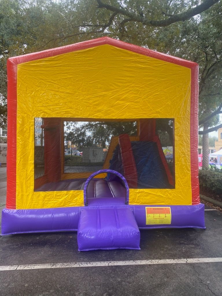A bright yellow, red, and purple inflatable bounce house with a small slide inside, set on an outdoor parking lot.