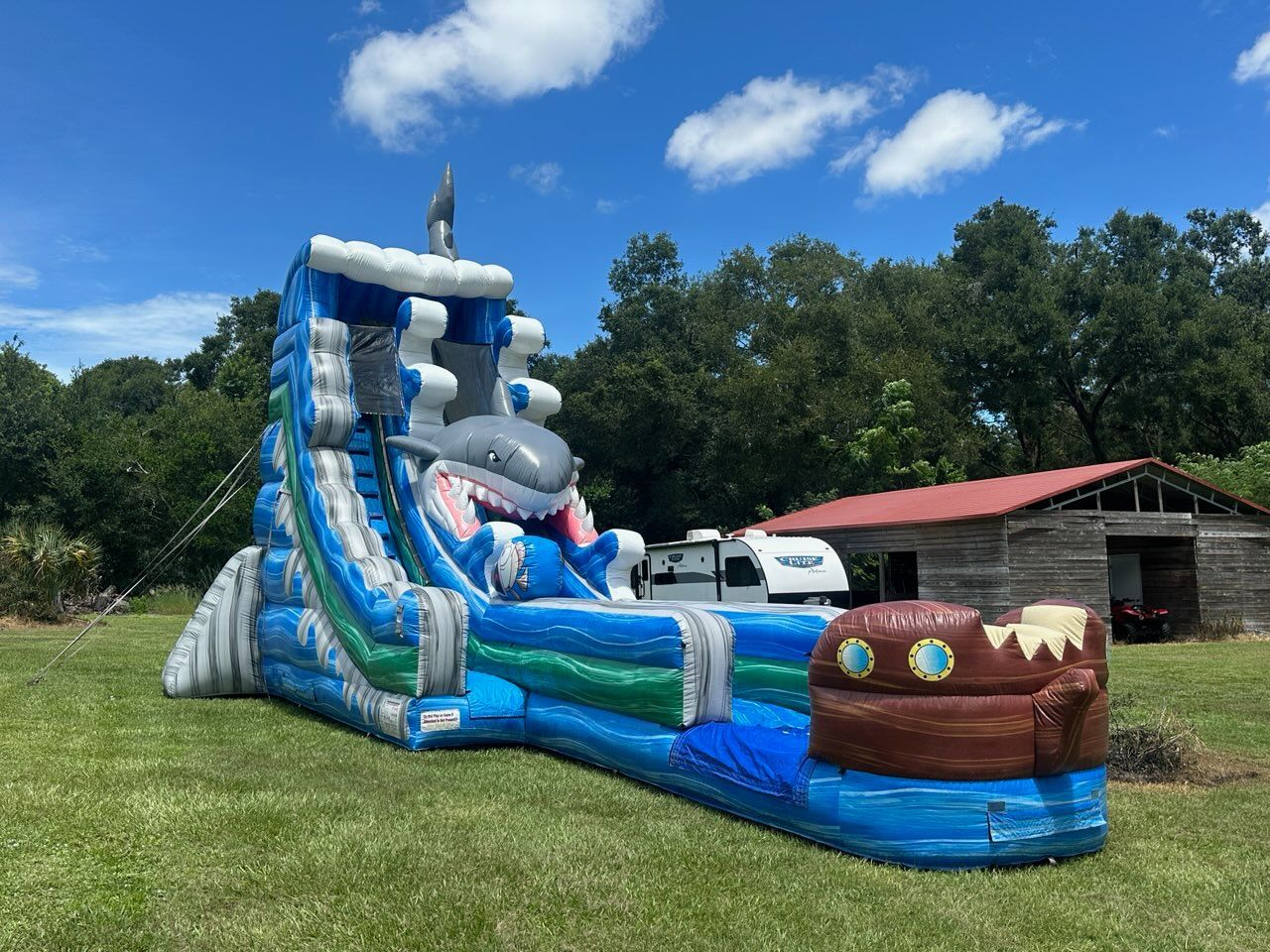 A blue and green inflatable shark water slide sits on a green lawn with a wooden building and blue sky in the background.