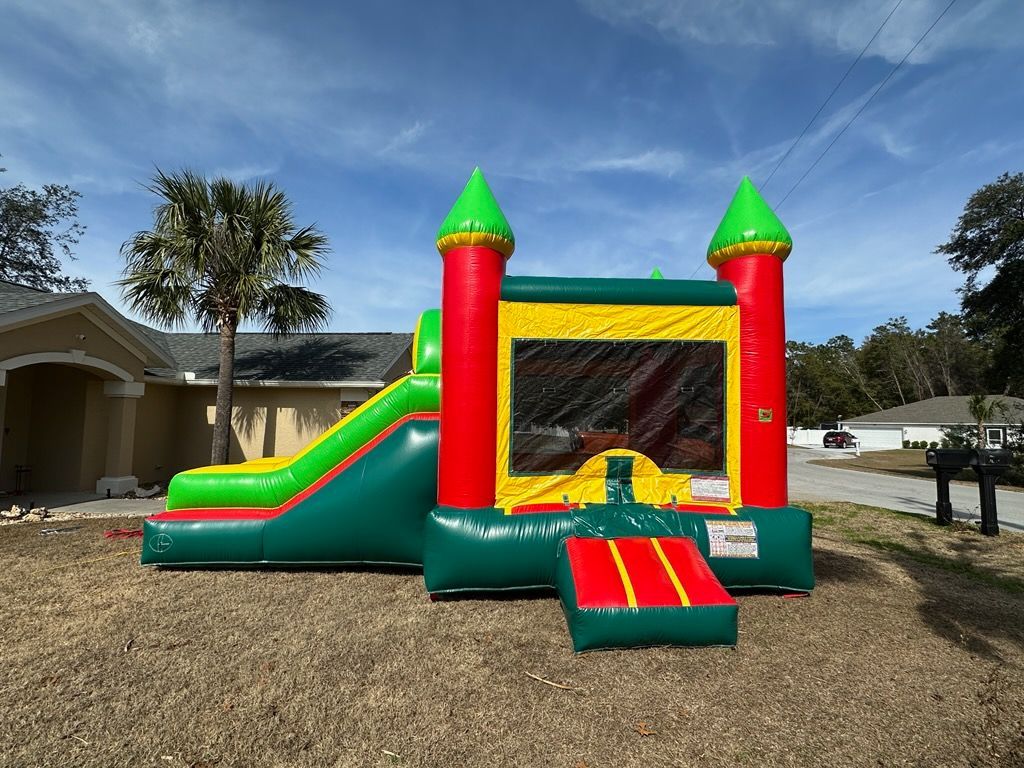 A colorful inflatable bounce house with a slide and two red and green towers, set on a dry grassy lawn outdoors.
