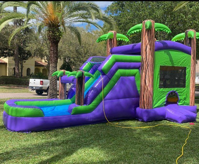 An inflatable tropical-themed bounce house with a slide and a pool area, set on a green lawn in front of trees and a house.