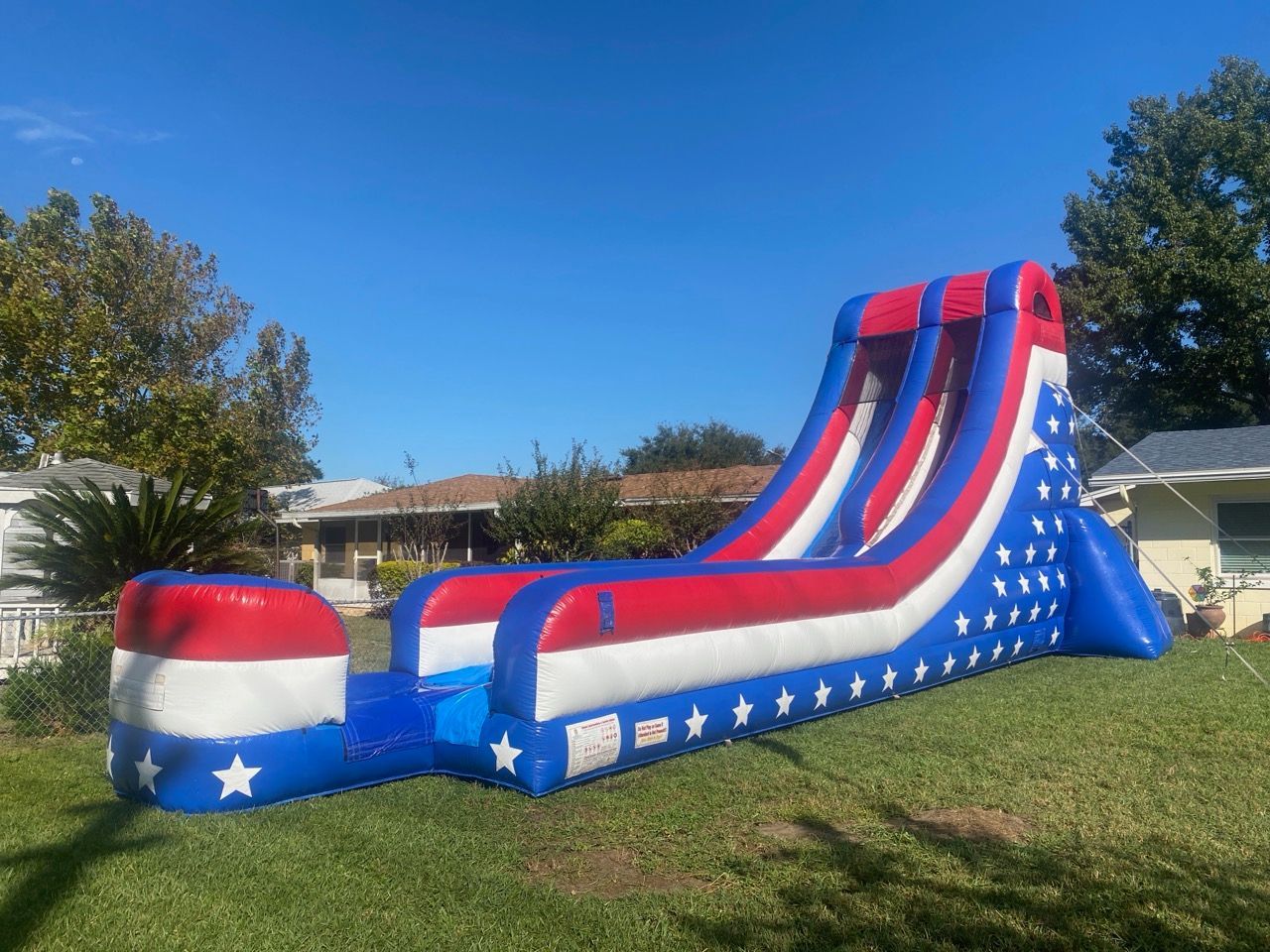 Inflatable red, white, and blue dual-lane water slide with star patterns, set on a residential lawn under a clear sky.