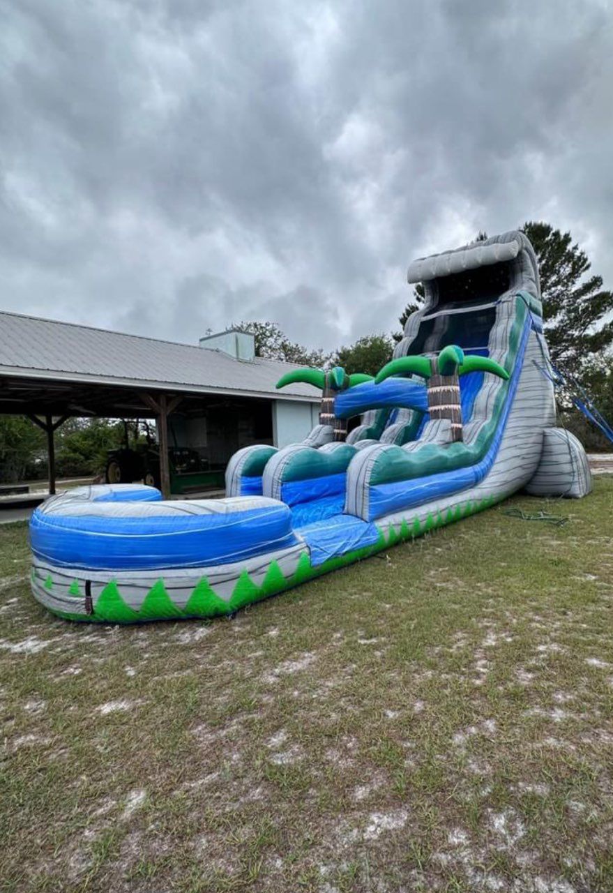 Inflatable water slide with palm tree decorations on a grassy field in front of a building under a cloudy sky.